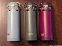 Front view of three Thermos water bottles in light gray, dark gray, and magenta on a wooden surface.