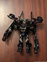 Front view of black Transformers Barricade action figure in robot form on a wooden surface showing detailed face, chest with police lights, arms with weapons, and wheels on shoulders.