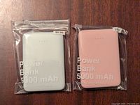 Two power banks packaged individually in clear resealable bags, placed on a wood surface. One is light blue, the other light pink. Both have 'Power Bank 5000 mAh' text on front and price stickers.