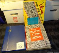 Two stamp books shown, one Majestic Stamp Album with vintage covers and another blue binder-style album along with Majestic Stamp Collectors Outfit box.