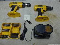 Two DeWalt cordless drills without batteries, a charger, and a drill bit set arranged on a beige surface.