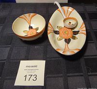 Overview of three pieces: large oval dish, ladle resting inside it, small bowl. All have orange flower design with gold and black leaves and orange fan accents. Placed on black textured cloth.