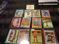 Top-down photo showing twelve 1970's hockey cards arrayed in rows on a black surface, including team photos and player portraits of Bobby Orr, Guy Lafleur, Bobby Hull, Ron Schock, and others.
