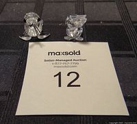 Two Swarovski crystal animal figurines, a seal and a pelican, displayed on black fabric with auction number card