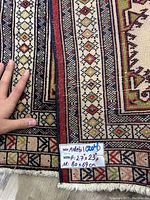 Close-up showing hand holding corner of rug with handwritten label indicating size 80x69 cm, handmade Ardebal rug.