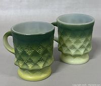 Two vintage Fire King Kimberly mugs shown side by side with gradient green to yellow coloring and diamond textured pattern.