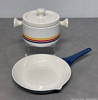 Blue handled cast iron frying pan with white interior alongside white ceramic pot with lid and rainbow stripes