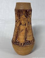 Full view of the tan ceramic vase with hand carved figures and textured top and bottom sections.