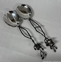 Two serving utensils: one spoon and one fork with hammered silver-tone bowls and blackened olive branch handles with detailed flowers and buds.