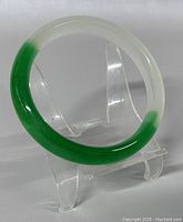 Natural jade bangle bracelet displayed on a clear stand, showing the green and white color gradient and polished surface.