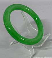 Bright green natural jade bangle displayed upright on clear acrylic stand, showing smooth rounded edges and glossy finish.