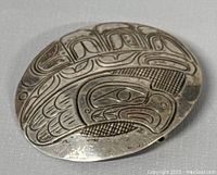 Front side of oval silver brooch with etched Indigenous Northwest Coast art design showing bird motifs.