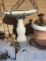 Full view of the assembled concrete bird bath standing against a brick wall with snow on the ground and adjacent plant pots.
