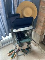 Full view of the garden tool stool with blue cushion, folded garden chair, black binoculars, and straw hat on top. Garden hand tools with wooden handles placed underneath.