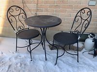 Photo of black metal bistro set showing round table with four curved legs and two matching chairs with swirl back designs seated on snow-covered ground.