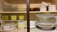 Kitchen cabinet shelf holding various vintage kitchenware items including Corelle plates, cereal bowls, Fire King casserole dishes with wheat pattern, and white ceramic ramekins.