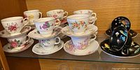 Fourteen teacup and saucer sets displayed including floral patterns in different colors and styles.