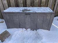 Front view of plastic deck box, showing closed lid with snow and visible damage on top