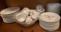 Full set of ceramic dinnerware including stacked dinner plates, side plates, bowls filled with cups, and grouped teacups with saucers on a wooden shelf.