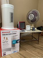 Photo showing Honeywell air purifier on its original box, Garrison ceramic heater, two Timex clocks, AcuRite humidistat, Super table fan on chrome metal frame table on casters.
