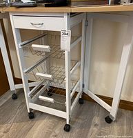 White folding stand with one drawer and three wire baskets, side panels folded inward showing hinge mechanism and caster wheels.