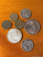 Top down view of seven foreign coins arranged on wood surface including a large Maria Theresa Thaler, Austrian Schilling, Mexican Pesos, French Francs, and Euro cents.