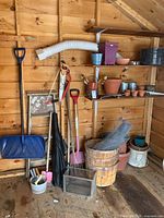 Wide angle photo showing gardening tools including shovels, rakes, pruners, a step ladder, enameled pot, clay pots, and baskets in a wooden shed