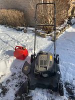 Overall front view of black Yardworks push lawn mower on a snowy surface with a red plastic gas container nearby.