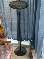 Full view of standing heater with black round base, adjustable stem, and heating element at the top in front of blue curtain.
