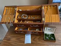 Open wooden jewellery box showing compartments with bracelets, earrings, brooches, and drawers open with jewelry inside on a wooden table.