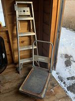 Five-foot aluminum step ladder and folding cart placed inside a shed near an open door with visible snow outside.
