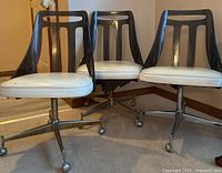 Three mid century dining chairs with smoked acrylic backs and white vinyl padded seats on metal caster bases, showing signs of wear and surface rust on metal parts.
