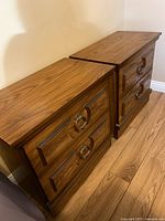 Pair of mid century night tables shown side by side with wood grain laminate tops and two drawers each featuring decorative metal ring handles