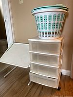 Storage unit with four semi-transparent drawers on caster wheels and foldable desk next to it with part of laundry baskets visible on top.