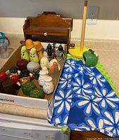 Full view of twelve assorted vintage salt and pepper shaker sets in a tray, wooden wall shelf behind, yellow paper towel holder to the right, and blue floral half aprons folded beside them.
