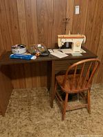 Full view of sewing machine on wood cabinet with chair, surrounded by sewing accessories including thread and tools.