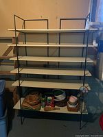 Full view of shelving unit showing six white shelves supported by black metal frame with various items on bottom shelf.