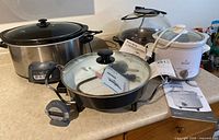 Four small kitchen appliances: a large unbranded slow cooker, an Oyster electric skillet with glass lid and user guide, a Rival 2-quart slow cooker with user guide, and a West Bend Stir Crazy Corn Popper with user guide, all placed on a kitchen counter.