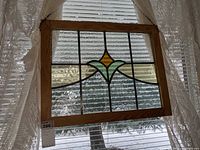 Clear view of the full stained glass panel with light wood frame, showing central colored design and textured clear glass background, hanging in front of window with lace curtains.