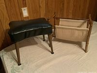 Photo of the mid century foot stool with black vinyl upholstery and four tapered wooden legs next to the bamboo framed magazine rack with canvas sling.