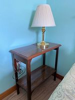 Full view of wood side table with single lower shelf and table lamp on top against blue wall.