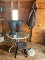 Patio set grouped inside wooden structure: round glass table, two plastic planters on table, brown umbrella folded, brown plastic side table, and white umbrella stand on floor.