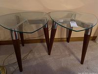 Pair of vintage style side tables with rounded triangular tempered glass tops, wood tapered legs, and chrome tripod frame separated on carpeted floor.