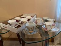 Wide angle showing the set of 5 Colclough teacups with matching saucers in a cardboard tray, 1 Royal Adderley teacup with intricate blue and gold floral design and no saucer, and 2 Roslyn China teacups with floral designs and gold trim, all arranged on a glass table.