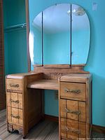 Front angle of vintage wood vanity showing tri-fold oval mirror and drawer arrangement with brass handles and wood finish.