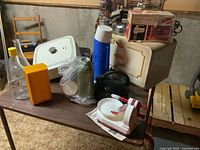 Vintage kitchen items arranged on a wooden table including electric frying pan, kettles, bread box, Thermos, apple peeler, bottles, and mixer