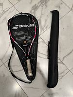 Full view of Babolat tennis racket with black, pink, and white frame inside its black carrying case, accompanied by black billiards cue case.