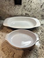 Photo showing a rectangular white ceramic casserole dish and a round dish with an orange rim on a countertop.