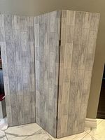 Three-panel folding room divider viewed standing on a floor with marble tiles, showing the grey wood plank pattern.