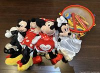 Four Mickey and Minnie Mouse plush toys arranged on floor: Mickey in black suit, Minnie in red polka dot dress, Minnie holding heart pillow with 'LOVE', Minnie in white wedding gown.